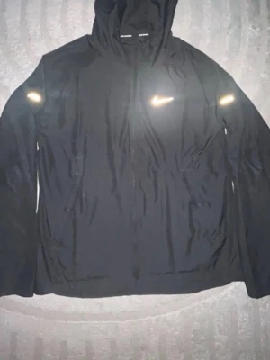 Svart Nike Miller - Säljer en svart Nike Miller från Nike med huva och dragkedja framtill. Jackan har reflekterande detaljer på bröstet och ärmarna, perfekt för löpning. Tillverkad i lätt material som andas och ger bra rörelsefrihet, perfekt för sommaren! PRIS KAN DISKUTERAS VID SNABB AFFÄR!