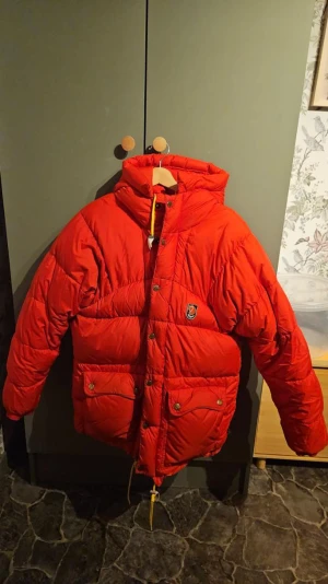 Röd Fjällräven Polar dunjacka XXS - Säljer en riktigt snygg röd dunjacka från Fjällräven Polar i storlek XXS. Jackan har huva, stora fickor med knappar och gula snören. Perfekt för kalla dagar och har den klassiska Fjällräven-loggan på bröstet. Finns någon liten fläck(bild). 
