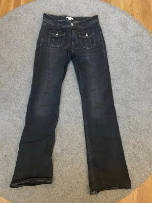 Svarta bootcut jeans Gina Tricot 170 - Snygga svarta bootcut jeans från Gina Tricot i storlek 170. Jeansen har två framfickor med lock och knapp, samt klassiska bakfickor. Tydliga kontrastsömmar och en lätt tvättad look ger dem en cool vibe. Perfekta till sneakers eller boots. Dom har använts men har i väldigt gott skick.