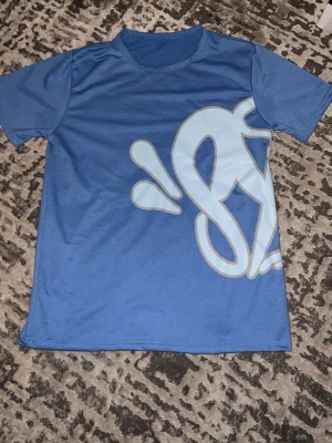 Blå Syna World t-shirt - Säljer en blå t-shirt med korta ärmar och stor ljusblå grafisk print som täcker både fram- och baksidan. T-shirten har rund hals och är gjord i ett mjukt syntetmaterial som känns skönt mot huden. Perfekt för dig som gillar streetwear och stora tryck.