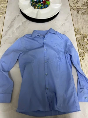 Ljusblå skjorta från M&S - Klassisk ljusblå skjorta från M&S med lång ärm och knappar framtill. Skjortan har en stilren krage och är tillverkad i ett mjukt bomullstyg. Perfekt för en clean och enkel look.