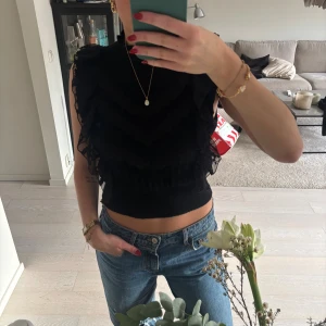 Svart spetsig croppad topp från Zara - Svart croppad topp från Zara med högt hals och breda volangdetaljer i spets längs axlar och bröst. Ribbad midja och ärmlös design ger en cool siluett. Perfekt för dig som gillar statement-plagg med mycket detaljer.