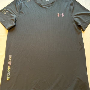 Svart Under Armour tränings-tshirt - Säljer en svart Under Armour t-shirt i polyester med korta ärmar och rund hals. Diskret logga på bröstet och färgglad UNDER ARMOUR-text längs sidan. Perfekt för löpning och träning, lätt och snabbtorkande material.