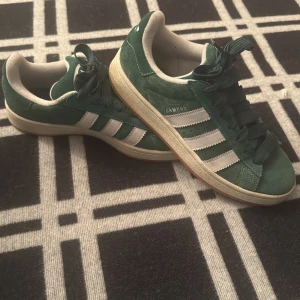 Adidas Campus gröna sneakers - Gröna Adidas Campus sneakers med klassiska vita ränder på sidorna och vit logga på hälen. Skorna har snörning, platt sula i gummi och är tillverkade i mocka med mjuk textil på insidan. Perfekt för dig som gillar retrostil och streetwear.