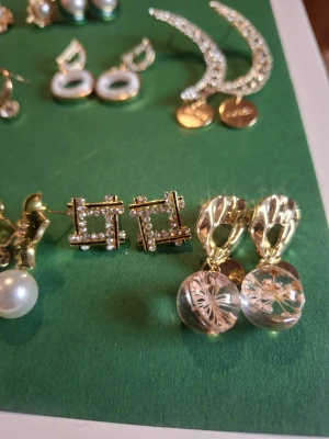 Mix av trendiga örhängen i olika stilar - Säljer ett set med flera par örhängen i olika former och färger. Här finns guldiga örhängen med pärlor, glittriga stenar, gröna blommor, lila blommor, hjärtan och cirklar. Vissa har detaljer som pärlor eller kristaller, andra är mer minimalistiska. Perfekt för dig som vill variera din look.