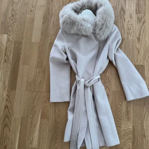 Hollies kappa med päls och bälte - Snygg beige kappa med stor fluffig päls runt kragen och knytbälte i midjan. Jackan har lång ärm och en stilren, färgen är ljusbeige. Behövs kemtvättas, därav pris