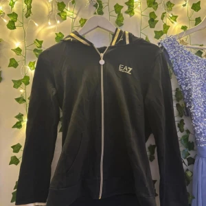 Svart zip-hoodie EA7 Emporio Armani - Svart hoodie med dragkedja från EA7 Emporio Armani. Snygga gula detaljer på huva och ärmslut med logotyp. Mjuk bomullskänsla, fickor fram och lång ärm. Perfekt för chill eller streetwear. Stilsäker look med märkets tryck på bröstet och huvan.