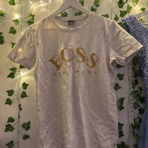 Vit t-shirt från Hugo Boss med guldtryck - Snygg vit t-shirt från Hugo Boss med stort BOSS-tryck i guld på bröstet. Klassisk rund hals och korta ärmar. Tillverkad i mjuk 100% bomull för skön känsla. Perfekt för dig som gillar stilrena märkesplagg med lite extra attityd.