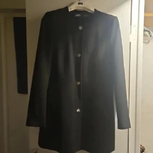 Svart rock från Zara med knappar - Stilren svart rock från Zara med raka linjer och guldfärgade knappar framtill. Jackan har lång ärm och enkel, klassisk design utan krage. Perfekt för dig som gillar minimalistisk stil och vill ha en snygg jacka till höst och vinter.