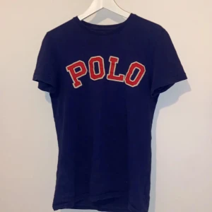 Blå Polo Ralph Lauren t-shirt - Snygg blå t-shirt från Polo Ralph Lauren med stor röd och vit POLO-text på bröstet. Modellen är kortärmad och har en klassisk rund hals. Passformen är custom fit och materialet är mjuk bomull, perfekt för en chill och stilren look.