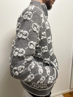 Grå stickad kofta från Gucci - Säljer en grå stickad kofta från Gucci med vit GG-logga över hela tröjan. Koftan har dragkedja framtill, ribbade muddar och hög krage. Den är långärmad och har fickor på sidorna. Perfekt för dig som vill ha en lyxig och snygg look.