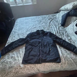 Svart vindjacka från Polo Ralph Lauren - Svart vindjacka från Polo Ralph Lauren med broderad logga på bröstet och stort P.R.L. Club-märke på ryggen. Jackan har dragkedja, två fickor med knappar och en broderad siffra 3 på ärmen. Snygg och sportig design, perfekt för dig som gillar klassisk streetstyle.