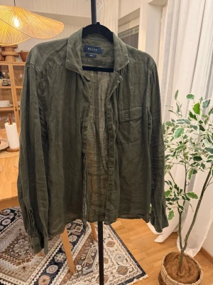 Olivgrön linneskjorta från Riley - Olivgrön skjorta från Riley i luftig linnemix, perfekt för dig som gillar avslappnad stil. Klassisk krage, knappstängning och en bröstficka med knapp. Långärmad modell med lite loose passform som ger en chill vibe.