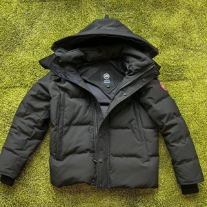 Svart pufferjacka från Canada Goose - Svart pufferjacka från Canada Goose med huva och klassisk röd logotyp på ärmen. Jackan har dragkedja och knappar framtill, ribbade muddar vid ärmslut och två sidofickor. Perfekt för kalla dagar med sin tjocka dunfyllning och stilrena look. Jackan har används 2 vintrar och därav pris och skick.