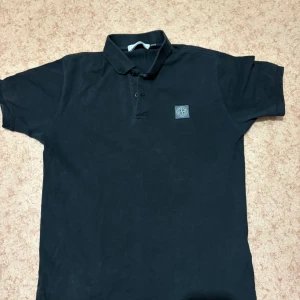 Svart pikétröja från Stone Island - Snygg svart pikétröja från Stone Island med klassisk krage och två knappar framtill. Tröjan har korta ärmar och den ikoniska Stone Island-loggan på bröstet. Perfekt för dig som gillar stilrena och sportiga plagg.