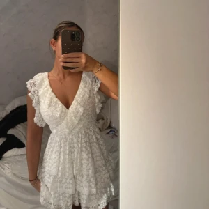 Vit spetsklänning med volanger - Supersöt vit kortklänning i spets med omlott och v-ringad front. Klänningen har volangdetaljer på ärmar och kjol, samt snörning i ryggen. Perfekt för sommarens alla tillfällen! Materialet är lätt och luftigt, och ger en romantisk vibe.