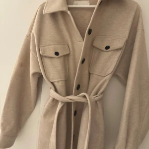 Beige kappa med bälte från nelly  - Snygg beige kappa från nelly, svarta knappar och ett matchande bälte i midjan. Jackan har lång ärm, klassisk krage och är gjord i mjukt syntetmaterial. Storlek xs men passar s. Har tyvärr en smink fläck kolla sista bilden