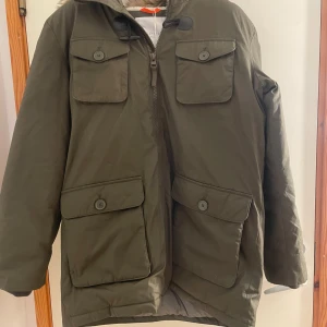 Olivgrön parkajacka från Esprit L - Parkajacka från Esprit i olivgrönt med fuskpäls på huvan, fyra stora fickor med lock och knappar, samt dragkedja framtill. Jackan är fodrad och har en loose passform, perfekt för kalla dagar. Materialet är syntet och polyester.