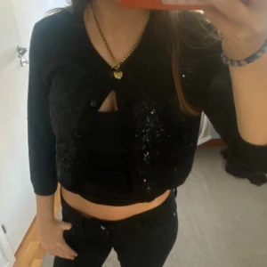 Svart paljettkofta från Sand Copenhagen - Snygg svart kofta från Sand Copenhagen med paljettdetaljer som ger extra glans. Koftan är croppad med trekvartsärm och knappar framtill. Perfekt för dig som vill sticka ut lite extra med en glittrig touch. Passar dig som gillar stilrena och trendiga plagg.