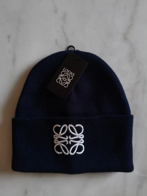 Loewe Beanie - Snygg mörkblå mössa från Loewe med vit broderad logga framtill. Klassisk uppvikt kant och mjukt material. Perfekt för dig som vill ha en clean och stilren look med exklusiv känsla. Taggar och etikett kvar.