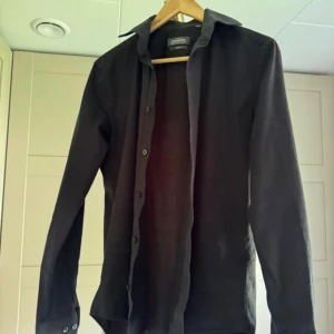 Svart slim fit skjorta Dressmann - Svart skjorta från Dressmann i slim fit-modell, tillverkad i stretchig bomull. Klassisk krage, långa ärmar och knappar hela vägen framtill. Perfekt för dig som gillar en stilren och enkel look. Storlek S 37/38