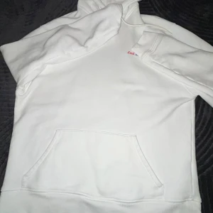 Vit hoodie från Axel Arigato - Vit hoodie från Axel Arigato med diskret logga i rött och blått på bröstet. Tröjan har huva, magficka och långa ärmar. Tillverkad i mjuk bomull för skön komfort och clean look. Perfekt för dig som gillar minimalistisk stil.