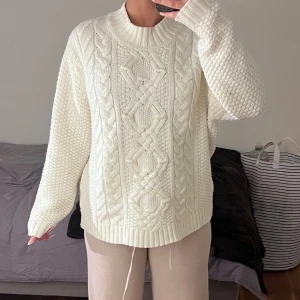 Vit stickad tröja med kabelmönster - Säljer en vit stickad tröja med grovt kabelmönster framtill och strukturstickade ärmar. Tröjan har rund hals och ribbade muddar vid ärmslut och nederkant. Perfekt för dig som gillar en oversized och mysig look.