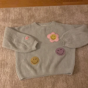 Ljusgrå stickad tröja med smileys - Supermysig ljusgrå stickad tröja med långärmad passform och ribbad struktur. Framsidan har broderade smiley-märken i gult och lila samt en rosa blomma med smiley. Perfekt för dig som gillar färgglada detaljer och chill vibe.
