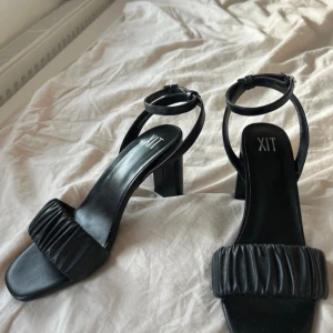 Svarta klackar  - Snygga svarta klackar från XIT med rynkad rem och justerbar ankelrem med spänne. Klacken är ca 9cm hög och passar 37-38. Perfekta för dig som gillar stilrena och trendiga skor.
