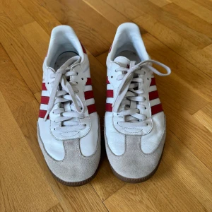 Adidas Samba OG sneakers vit/burgundy strl 44⅔ - Adidas Samba sneakers i vit och burgundy färg med de ikoniska tre ränderna på sidan. Ovandel i skinn med mockadetaljer på tån och gummisula. Snygg retrostil med snörning och rund tå, perfekt för dig som gillar streetwear och sportig look.