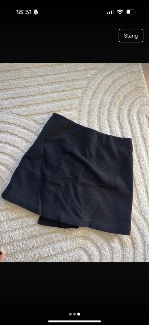 Svart omlottkjol från Stylein - Stilren svart kortkjol från Stylein i omlottmodell. Kjolen har en enkel och clean look med rak passform och är tillverkad i ett mjukt tyg som känns lite som ull. Perfekt att matcha med boots eller sneakers för en cool vibe.