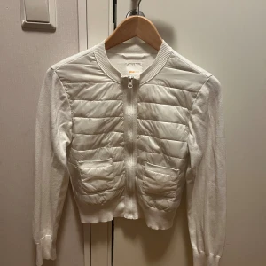 Vit quiltad cardigan jacka Gina Tricot - Snygg vit cardigan jacka från Gina Tricot med quiltad front och två fickor. Jackan har ribbad krage och muddar, dragkedja framtill och långärmade stickade ärmar. Perfekt för dig som gillar en clean och fräsch look.