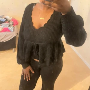 Svart stickad tröja med knyt - Svart stickad tröja med scallop v-ringad hals och långa ärmar. Tröjan har en snygg peplum-effekt med knyt i midjan och ribbad struktur. Perfekt för dig som gillar en feminin och trendig look. Som ny skick,knappt använd.