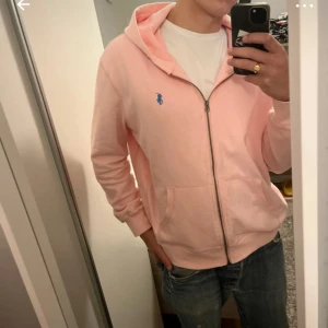 Rosa hoodie från Polo Ralph Lauren - Snygg ljusrosa hoodie från Polo Ralph Lauren med blå logga på bröstet. Tröjan har huva, dragkedja framtill och två fickor. Mjuk bomullskvalitet och ribbade muddar vid ärmslut och nederkant. Perfekt för chill dagar.
