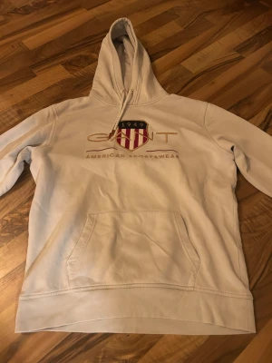Vit hoodie från GANT med broderi - Vit hoodie från GANT med broderat logotyp och sköld på bröstet. Tröjan har huva med snörning, känguruficka och ribbade muddar. Materialet är mjuk bomull och passformen är normal. Perfekt för dig som gillar klassisk amerikansk sportstil.