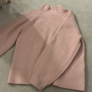 Ljusrosa ribbad stickad tröja - Superfin ljusrosa stickad tröja med ribbad struktur och hög hals. Tröjan har långärmade ärmar och en relaxed fit som är både snygg och bekväm. Perfekt att styla med jeans eller kjol för en chill look.