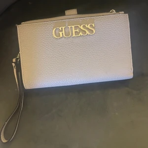Beige plånbok från Guess med dragkedja - Snygg beige plånbok från Guess med guldigt logotyp på framsidan. Plånboken har flera kortfack, fönster för ID, dragkedjefack och avtagbar handledsrem. Tillverkad i strukturerat syntetmaterial som ger en lyxig känsla.