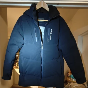 Mörkblå Sail Racing dunjacka - Säljer en äkta mörkblå dunjacka från Sail Racing (se tag på bilden), modell Jr Patrik down jacket, storlek S/170 cm. Jackan har huva, två bröstfickor med dragkedja och snygga Sail Racing-loggor på bröstet och ärmen. Perfekt för kalla dagar med sin tjocka och värmande design. Det finns två jack, ett under tejpen och ett på baksidan (se bilder) utöver det så är den i helt nytt skick.