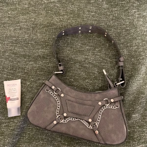 Svart axelväska med kedjedetaljer - Cool liten grå axelväska i syntetmaterial med  kedjor och nitar. Väskan har en någorlunda justerbar rem och stängs med dragkedja. Säljer då den aldrig kommer till användning