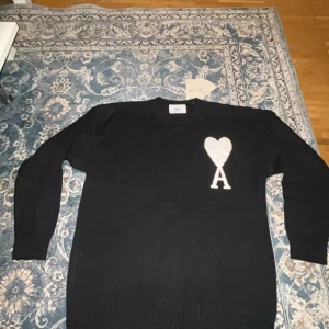 Svart stickad tröja från Ami Paris - Svart stickad tröja från Ami Paris med vit hjärtapplikation och bokstaven A på bröstet. Tröjan har rund hals, ribbade muddar och lång ärm. Tillverkad i Portugal av mjuk ull, perfekt för att hålla dig varm och snygg under kyliga dagar.
