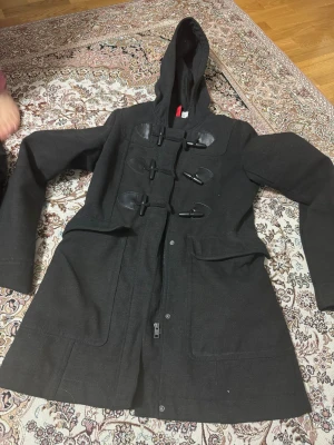 Svart rock med huva från Divided - Svart rock från Divided med huva och snygga snörningsdetaljer framtill. Jackan har långa ärmar, stora fickor och dragkedja. Yttermaterialet är ullblandning och insidan är fodrad med syntet. Perfekt för kalla dagar när du vill ha en stilren look.