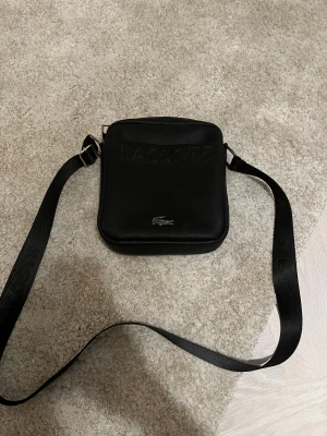 Svart axelväska från Lacoste - Snygg svart axelväska från Lacoste med justerbar rem och diskret logga framtill. Väskan är tillverkad i slitstark syntet med en stilren, fyrkantig form och dragkedja upptill. Perfekt för att bära det viktigaste med stil. Säljer för det är inte min stil. Dragkedjan kan fastna då och då.