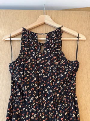 Svart långklänning med blommigt mönster - Supersnygg svart långklänning från Tom Tailor med små blommor i beige, rött och orange. Klänningen har ärmlös halterneck, öppen rygg med snygg detalj och elastisk midja för skön passform. Perfekt för dig som gillar boho-vibe och mönstrat. Mönstrad halterneck maxiklänning, märke Tom Tailor Denim köpt på Zalando. Perfekt till tex nyår eller vardags med tröja för höst vinter vajb.