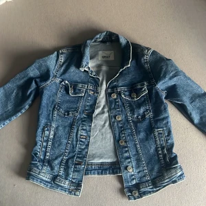 Blå jeansjacka från ONLY - Snygg klassisk blå jeansjacka från ONLY med silverfärgade knappar och flera fickor fram. Jackan har lång ärm, krage och är tillverkad i slitstarkt jeanstyg. Perfekt att slänga över en hoodie eller t-shirt.