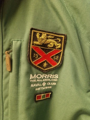 Grön jacka från Morris i Small - Snygg grön jacka från Morris med broderad logga på bröstet i storlek Small. Fodret har ett stort brittiskt flaggmotiv som ger extra karaktär. Lite mörkare grön än vad det är på bilderna på hela jackan, se detaljbilden. 