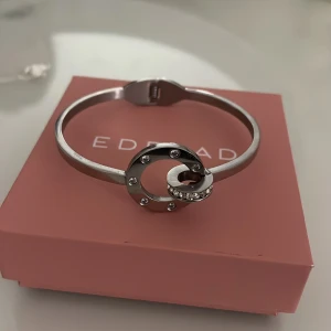 Silverfärgat armband från Edblad - Stilrent armband i silverfärg från Edblad med cirkelformad detalj framtill, dekorerad med små gnistrande stenar. Armbandet har en smal, stel design och ett smidigt lås. Perfekt för dig som gillar minimalistiska smycken med lite extra bling. Kan tänka mig gå ner i pris men just nu är den hälften av originalpriset.