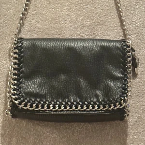 Väska - Svart axelremsväska i skinnimitation kantad med silverfärgad kedja. Ett fack med dragkedja på baksidan och ett fack med dragkedja på insidan. Axellänk 105 cm. Längd 28 cm, höjd 18 cm, bredd 5 cm.