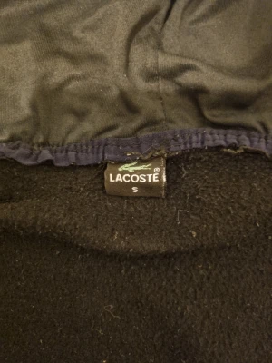 Svart fleecejacka med huva från Lacoste - Svart fleecejacka från Lacoste i storlek S. Jackan har huva, hel dragkedja och två fickor med dragkedja. Paneler i syntetmaterial på bröstet och detaljer med Lacoste-logga. Perfekt för dig som gillar sportig och clean stil.