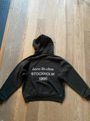 Svart hoodie Acne Studios Stockholm - Svart hoodie från Acne Studios med stor vit text på ryggen: 'Acne Studios STOCKHOLM 1996'. Klassisk passform med huva och långa ärmar. Mjuk bomullskvalitet. Perfekt för dig som gillar streetwear och enkel stil.