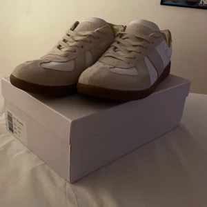 Maison Margiela sneakers i vitt och beige - Maison Margiela sneakers med vit och beige ovandel i skinn och mocka, klassisk rund tå och platt gummisula i brunt. Snygga paneler och diskret branding med ikoniska siffror på sula och låda. Perfekt för dig som gillar minimalistisk och stilren design.